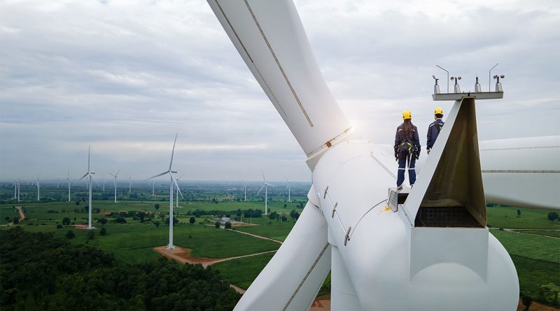 Two engineers in safety gear standing atop a wind turbine nacelle, overlooking a large wind farm spread across green fields under a cloudy sky.