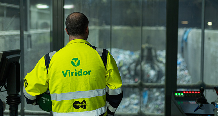a person wearing a high-visibility jacket with the "Viridor" logo on the back, standing in a control room and looking out at a large industrial facility