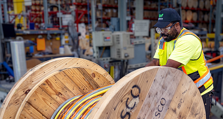 a man in a warehouse or industrial setting, wearing a yellow high-visibility vest, a black cap with a "W" logo, and safety glasses. He is working with two large wooden spools of multi-colored electrical or data cables